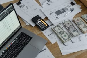 Mesa com laptop mostrando planilha, calculadora, notas de dinheiro e documentos financeiros