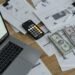 Mesa com laptop mostrando planilha, calculadora, notas de dinheiro e documentos financeiros
